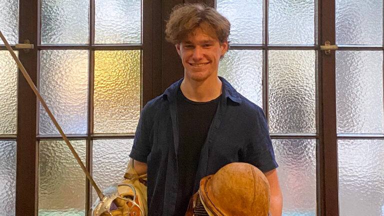 Student Vladimir (25) mit einem Teil seiner Ausrüstung zum studentischen Fechten vor einem Fenster. In den Händen hält er einen Helm und seine scharfe Klingenwaffe.