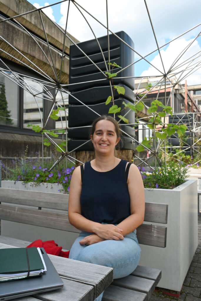 Anna Carmen Breuer, erste Klimaanpassungsmanagerin der Hochschule Darmstadt, sitzt auf einer Bank vor einem modernen Gebäude mit Metallstrukturen und Pflanzen