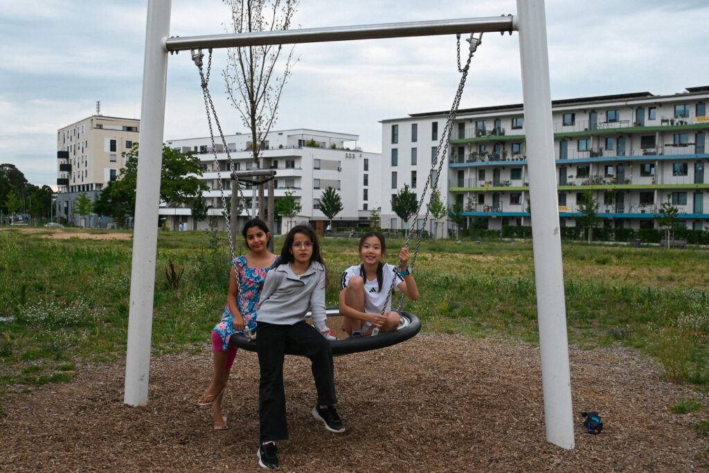 Eleni B. sitzt in der Mitte mit zwei Freundinnen auf einer runden Schaukel im Quartierspark der Lincoln-Siedlung, umgeben von modernen Wohngebäuden